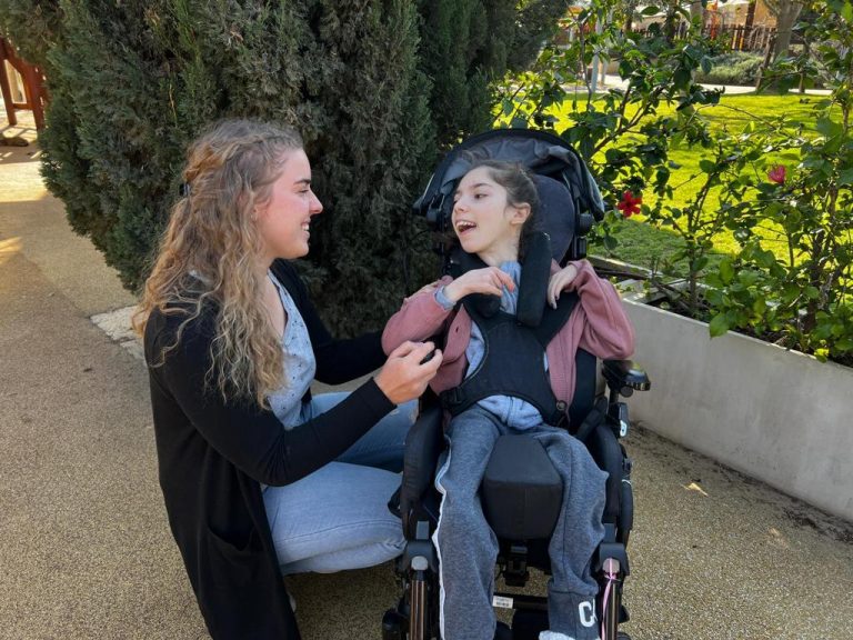 ילדה עם מוגבלויות בכסא גלגלים מחייכת למתנדבת Girl with disabilities in a wheelchair smiling at volunteer